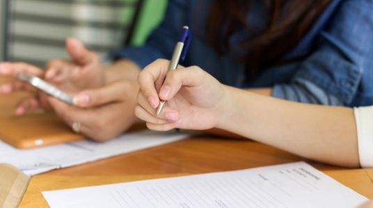 Two people's hands. One holds a pen, another holds a calculator.